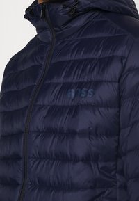 Mörkblå puffjacka med dragkedja, featuring horisontellt quiltat mönster och justerbar huva. Litet präglat logotyp på bröstet.
