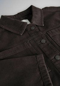 Veste en denim marron foncé avec un col pointu, des boutons-pression en métal et des poches poitrine visibles sur une surface plane.