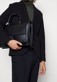 Svart läderportfölj med dubbla handtag, slät struktur och framficka. Matchad med en mörk blazer och grön polotröja.