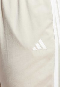 Svetlobéžové športové nohavice vyrobené z mäkkej látky, s tromi bielymi pruhmi na bokoch a bielym vyšívaným logom Adidas.