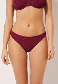 Burgunderfarbene Bikinihose mit strukturiertem Muster, mittelhohem Schnitt und glatter Oberfläche. Sie zeichnet sich durch ein minimalistisches Design und dezente seitliche Akzente aus.