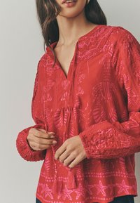 Blusa rossa con motivi floreali e stelle rosa, dotata di scollatura e maniche elasticizzate, oltre a lacci con pon-pon nella parte anteriore.