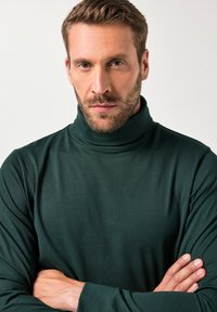 Tumma vihreä poolopaita, joka on valmistettu sileästä, kevyestä kankaasta ja jossa on korkea kaulus sekä pitkät hihat, joissa on tiukka muotoilu.