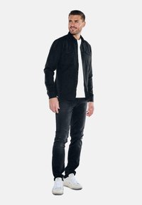Schwarze Jeansjacke mit Knopfverschluss, zwei Brusttaschen, getragen über einem weißen T-Shirt, kombiniert mit dunkelblauen Jeans und weißen Sneakers.
