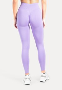 Lila Leggings mit hoher Taille aus glattem, dehnbarem Stoff, mit einer konturierten Rückennaht zur Formverbesserung. Kombiniert mit weißen Turnschuhen.