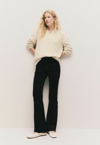 Cremfarbener, strukturierter, übergroßer Pullover, schwarze Cordflared-Hosen und hellfarbene Ballerinas, getragen von einer Person, die vor einem neutralen Hintergrund posiert.