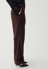 Smeđe krojene hlače ravnog kroja, s detaljima na džepovima i zakovicama na rubu. Nosi se s crnim cipelama s oštrim vrhom.