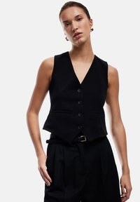 Schwarze Weste mit tiefem V-Ausschnitt, Vorderknopfleiste und zwei Taschen. Kombiniert mit hochtaillierten schwarzen Hosen, die einen Gürtel mit einer goldenen Schnalle haben.