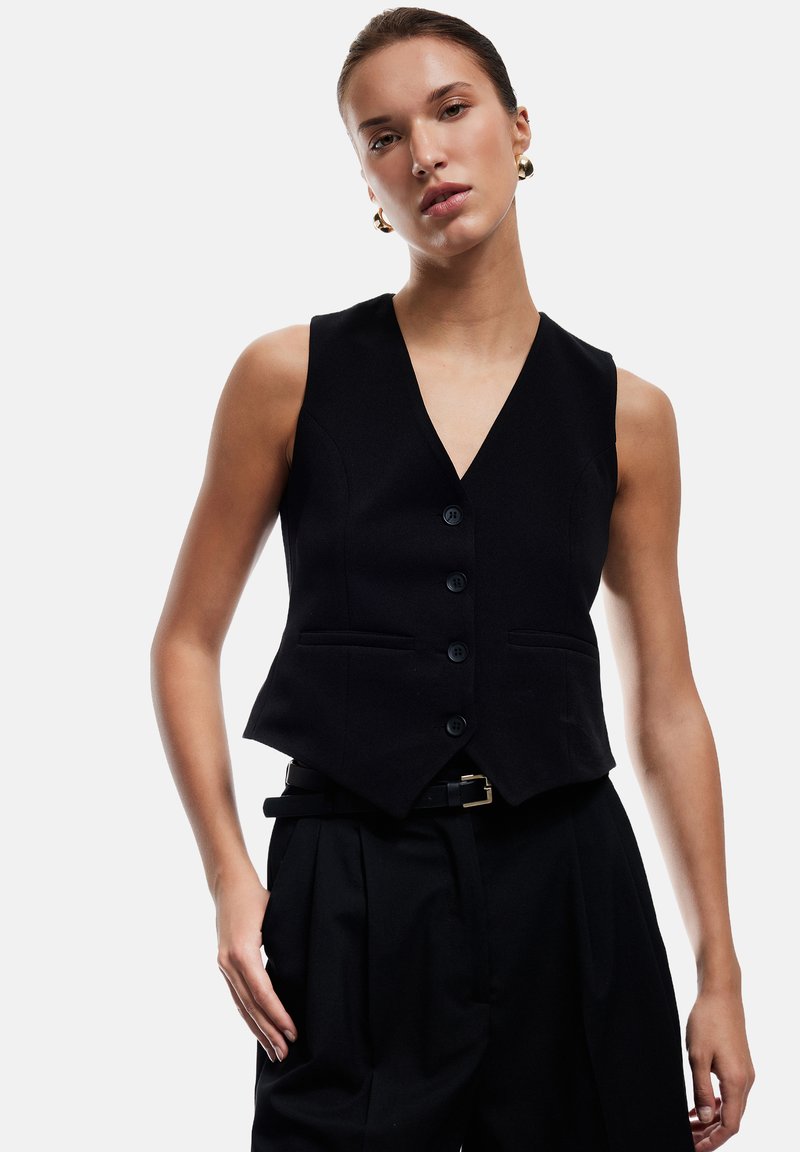 Chaleco negro con un escote en V profundo, cierre frontal de botones y dos bolsillos. Superpuesto a unos pantalones negros de cintura alta, que cuentan con un cinturón con hebilla dorada.