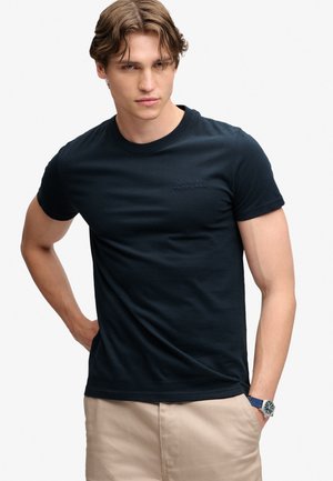 Junger Mann, der ein eng anliegendes schwarzes T-Shirt und beige Hosen trägt, mit einer Hand in der Tasche und kurzen welligen Haaren, stehend vor einem schlichten Hintergrund.