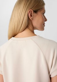 Lichtbeige top van zachte, soepele stof. Heeft een ronde halslijn, korte mouwen en een subtiele geborduurde logo op de achterkant.