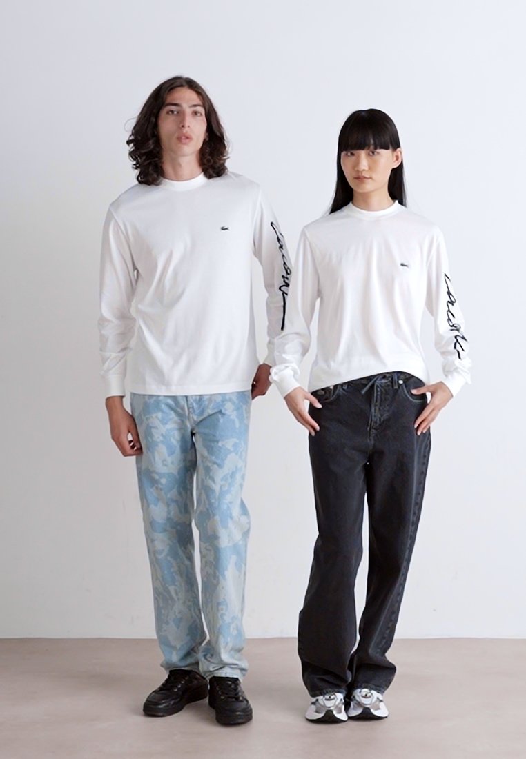 Chemises blanches à manches longues avec de petits logos sur la poitrine, associées à un camouflage bleu et à des jeans noirs, présentées par deux modèles.