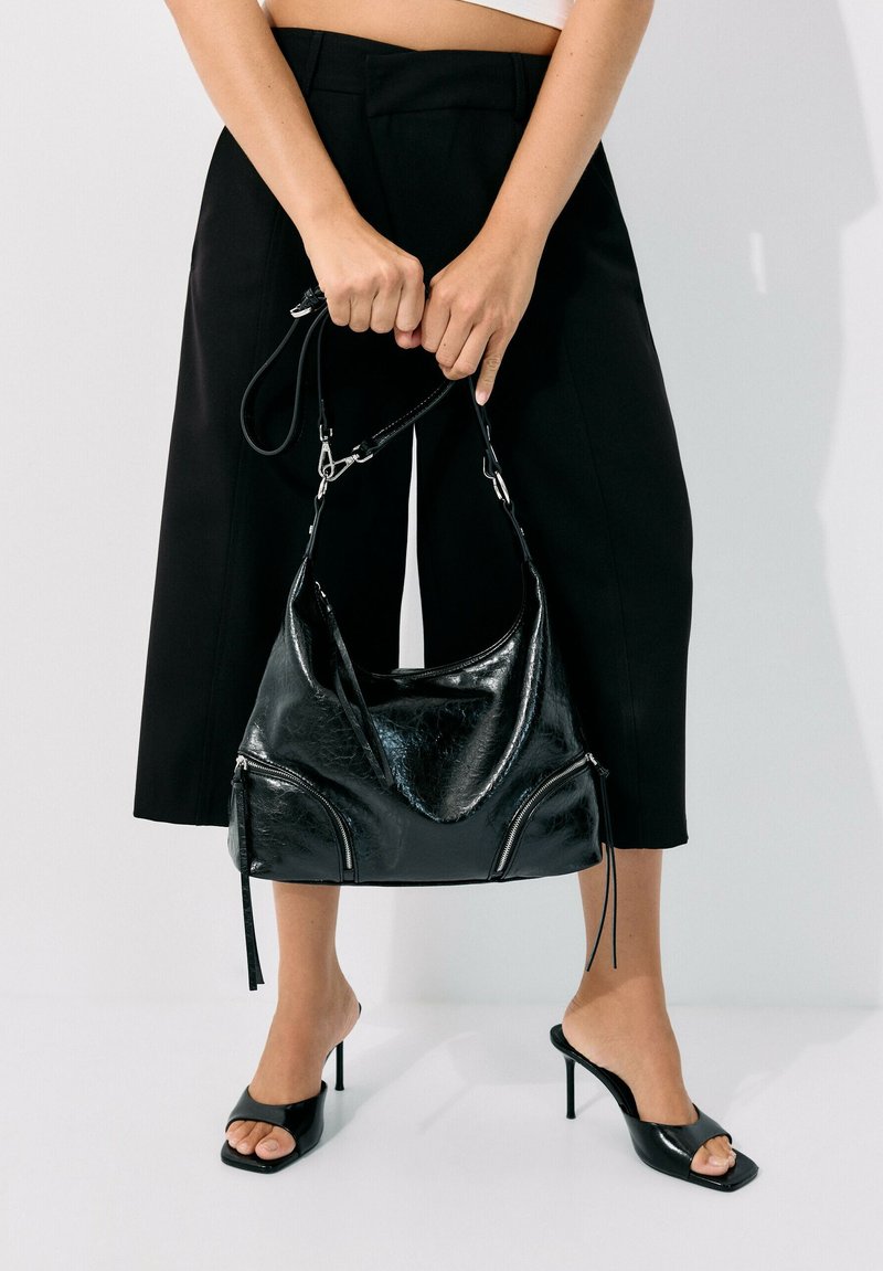 Personne portant un pantalon noir à jambes larges et des sandales noires à talons hauts tenant un sac à main en cuir noir brillant par ses anses.