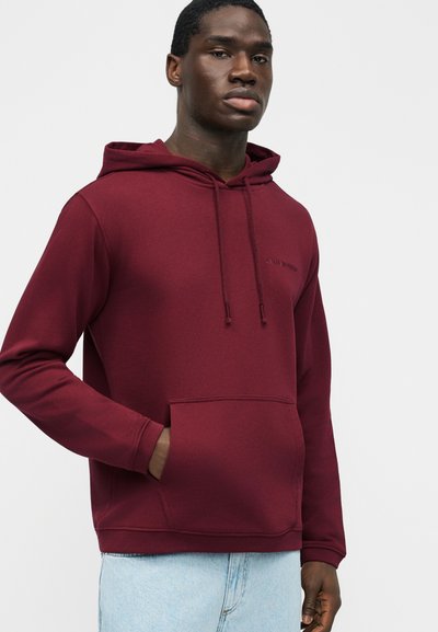 Άνδρας που φοράει μπορντό φούτερ με κουκούλα και μπροστινή τσέπη, καθώς και ανοιχτό μπλε τζιν παντελόνι, στέκεται μπροστά σε μια απλή ανοιχτή επιφάνεια.