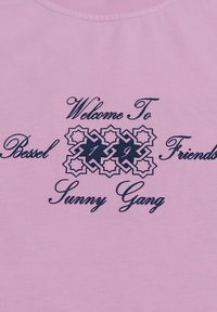 Camiseta de algodón rosa con texto impreso en azul marino: "Bienvenido a Basilea 199 Amigos Sunny Gang", con detalles de patrón decorativos.