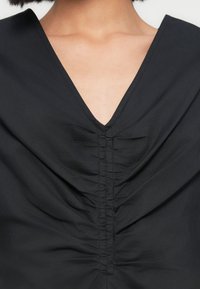 Μαύρο τοπ με βαθύ V-λαιμόκοψη, με λεπτομέρειες πτυχώσεων στην μπροστινή πλευρά, κατασκευασμένο από απαλό ύφασμα με μαλακή υφή.