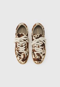 Ζευγάρι αθλητικά παπούτσια χαμηλού προφίλ με σχέδιο από δέρμα αγελάδας σε καφέ και λευκό και μπεζ κορδόνια, φαίνονται από πάνω σε απλό φόντο.