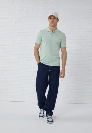 Tricou polo verde deschis cu mâneci scurte, pantaloni lungi largi de culoare bleumarin și o șapcă albă. Adidașii au un amestec de accente albastre închise și deschise.