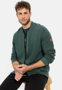 Dunkelgrüne Zip-Jacke aus strukturiertem Material, mit gerippten Bündchen und einem Lederlogo-Patch am linken Ärmel. Über einem schwarzen T-Shirt getragen.