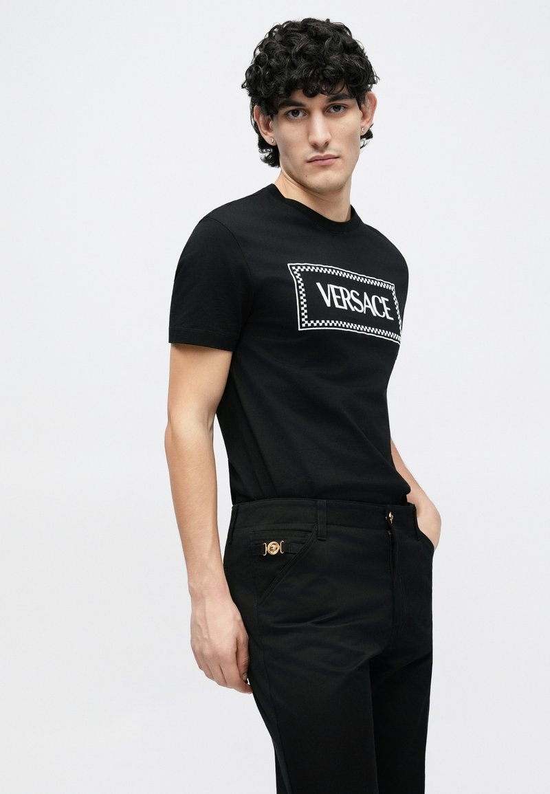 Svart t-shirt med korta ärmar som har en vit "VERSACE"-logotyp inom en rektangulär ram med ett rutmönster. Kombinerad med svarta byxor.