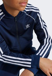 Mornarsko modra Adidas trenirka z belimi črtami na rokavih, z zadrgo spredaj in rebrastimi manšetami. Ima logo na prsih in gladko tekstilno površino.