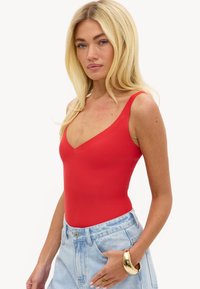Femme blonde portant un haut rouge sans manches et un jean bleu clair avec un bracelet et une bague en or, posant devant un fond blanc.