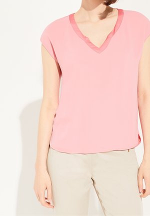 Kobieta w jasnoróżowej bluzce z krótkim rękawem i dekoltem w serek, w połączeniu z beżowymi spodniami, stojąca na białym tle.