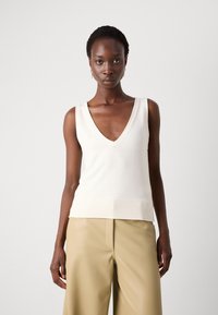 Top en tricot sans manches à col en V de couleur crème, présentant un ourlet côtelé, une texture douce et une coupe ajustée, associé à un pantalon large de couleur taupe clair.
