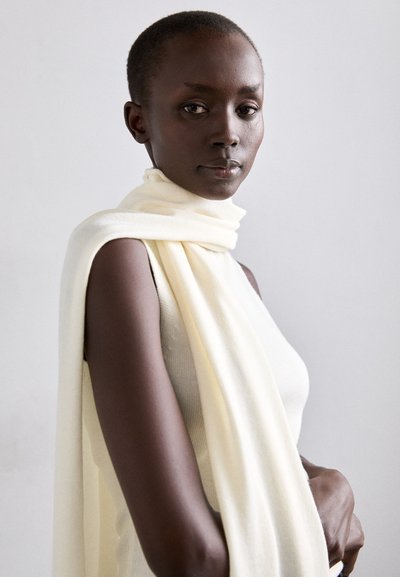 Écharpe en tricot de couleur crème drapée sur un haut sans manches beige clair, mettant en valeur sa texture douce et son design minimaliste sur un fond neutre.