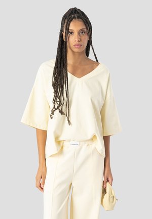 Mujer con cabello largo trenzado, vistiendo una blusa suelta color crema con escote en V y pantalones a juego, sosteniendo un bolso pequeño color crema sobre un fondo liso.