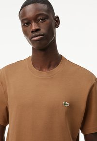 T-shirt in cotone marrone con scollatura rotonda e piccolo logo ricamato in verde e bianco sul lato sinistro del petto. Texture liscia.