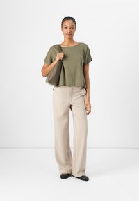 Maglietta verde oliva a maniche corte, pantaloni beige a gamba larga e ballerine nere. La persona tiene in mano una borsa a tracolla marrone. Tessuto morbido, vestibilità rilassata.