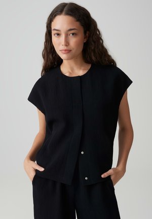 Vrouw met lang krullend haar draagt een losse zwarte top met korte mouwen en bijpassende broek, staand met handen in de zakken tegen een lichte achtergrond.