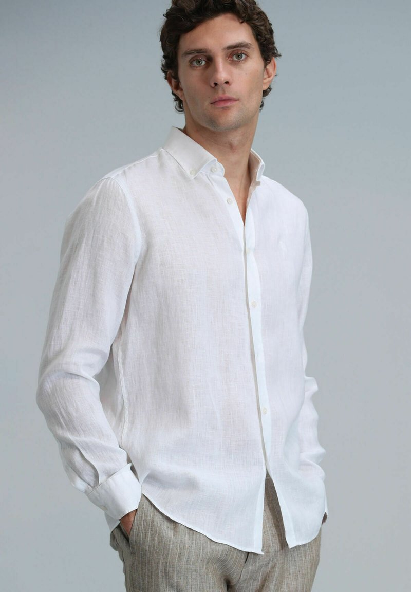Chemise blanche en lin à manches longues avec col boutonné, présentant une coupe décontractée et une texture subtile. Associée à un pantalon rayé beige clair.