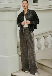 Zwarte leren bomberjack, beige top en grijze corduroy wijde broek met een trekkoord in de taille; model staat tegen een kolom.