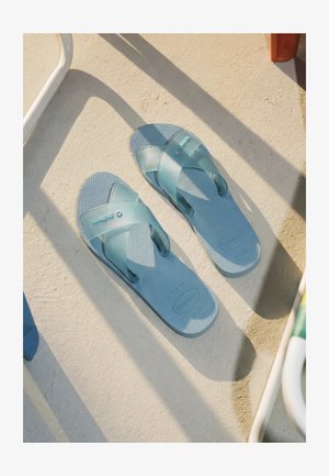 Paar doorschijnende blauwe Havaianas-sandalen op een gestructureerde beige vloer met zonlicht en schaduwen van nabijgelegen objecten.