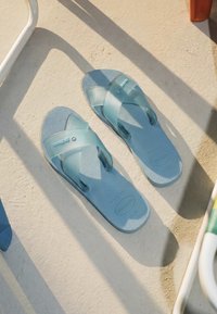 Paire de sandales Havaianas bleues translucides sur un sol beige texturé avec lumière du soleil et ombres d’objets à proximité.