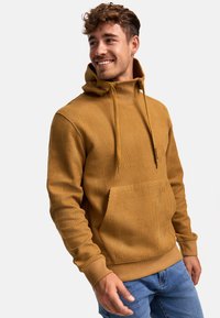 Sweat à capuche moutarde avec un motif texturé, cordons de serrage réglables et une poche frontale. Poignets et ourlet côtelés. Porté avec un jean bleu.