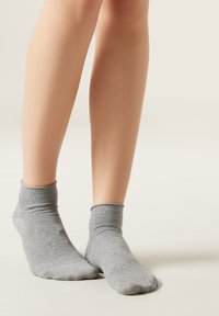 Calcetines tobilleros de algodón gris con un ribete de canalé, que muestran una textura suave y un diseño ligero, posicionados sobre piernas al descubierto contra un fondo neutro.