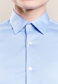 Närbild av en person som bär en ljusblå skjorta med spetsig krage och tre vita knappar knäppta.