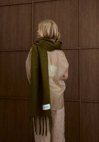 Écharpe en maille vert olive avec des bords frangés, portée sur un haut léger beige translucide, avec une étiquette marquée "ARCTIC FOX". Fond en bois.