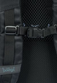 Zaino in rete nera con dettagli come spallacci regolabili, una fibbia in plastica e un'etichetta con marchio. Superficie testurizzata con design in rete traspirante.