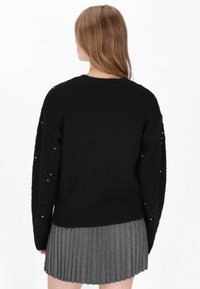 Fekete kötött pulóver, texturált fekete ujjakkal, amelyek flitterekkel díszítettek, szürke redőzött szoknyával párosítva. A hátsó nézet bemutatja a tervezési részleteket.