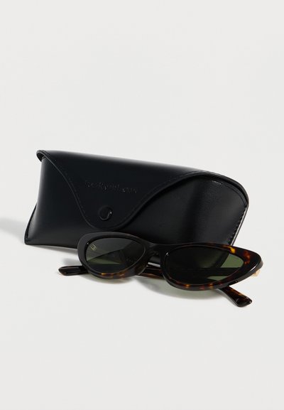 Γυαλιά ηλίου με σκελετό «μάτια γάτας» σε χρώμα χελώνας και σκουρούς φακούς, τοποθετημένα μπροστά από μια μαύρη δερμάτινη θήκη με κούμπωμα κουμπί.