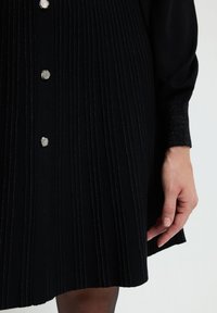 Abito nero con pieghe verticali, fili metallici e accessori con bottoni argentati, con maniche lunghe e un tessuto texturizzato.