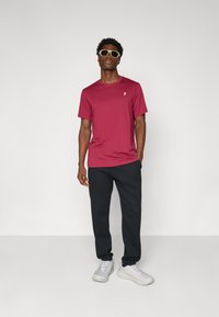 Maroon short-sleeve t-shirt with a small white logo, paired with black sweatpants and light gray sneakers. Model wearing sunglasses.