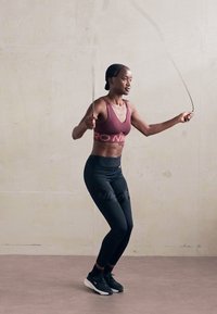 Sportos nő ugrál egy kötélen a teremben, bordó sportmelltartót és fekete leggingset viselve, miközben a edzésére összpontosít.