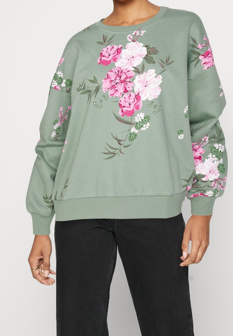 Sweat-shirt vert doux avec des imprimés floraux roses et blancs sur le devant et les manches, porté avec un pantalon noir, montré du cou aux hanches.