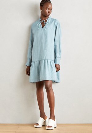 Rochie din in bleu deschis, cu o margine în trepte, decolteu legat și mâneci lungi. Pur și simplu cu mule albe cu toc bloc. Textură netedă.