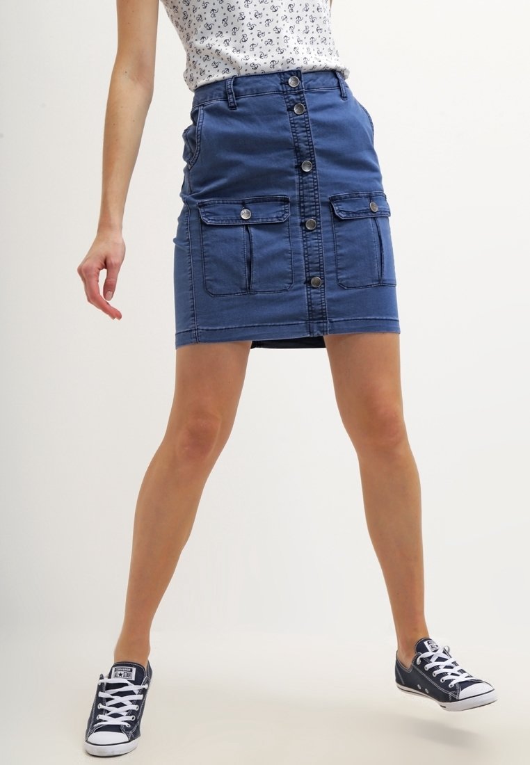 Mujer vestida con una falda de mezclilla azul con botones en la parte frontal, bolsillos con solapa y zapatillas negras de corte bajo, de pie con las piernas separadas contra un fondo blanco.